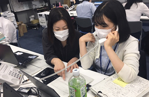 オフィス内で電話をとっている風景写真(人は皆、マスクをつけている)。